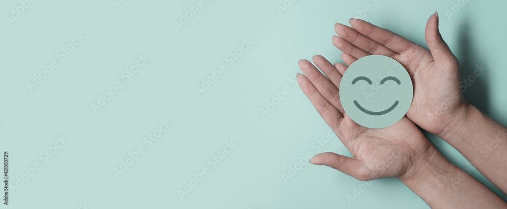 Image of a light blue background with two hands holding a blue smiley face made of paper.