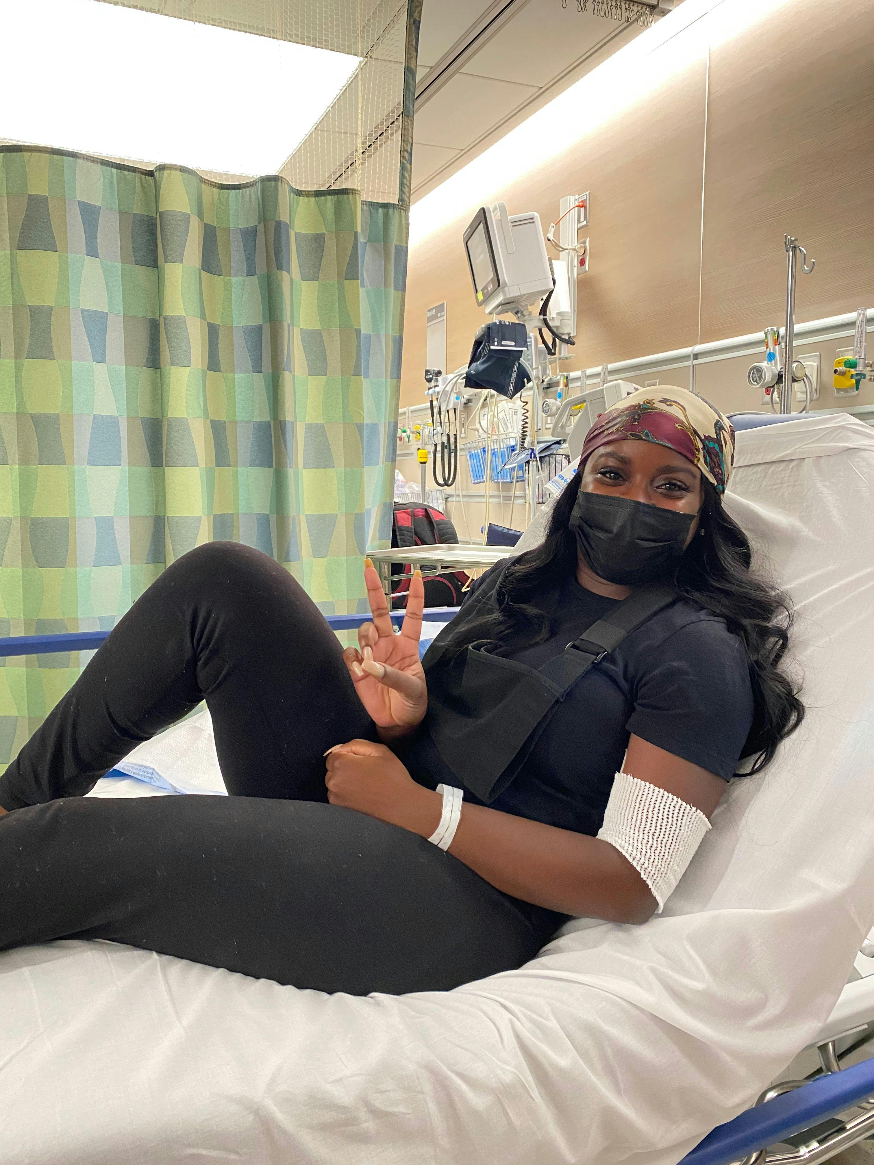 Paula Ngon, a black woman wearing a headscarf and mask, sits on a hospital bed, giving the "peace" sing | Photo provided by Paula Ngon
