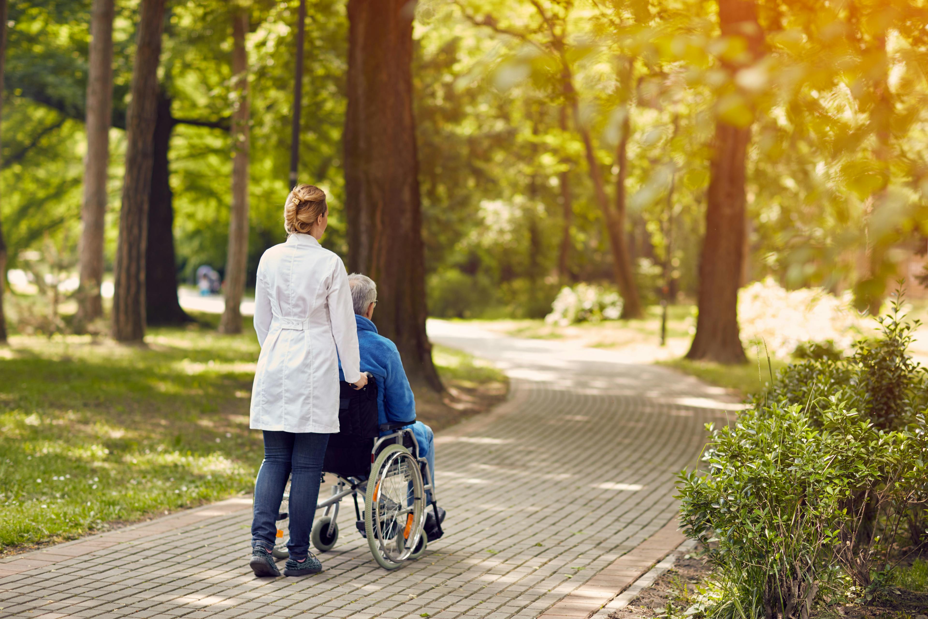 Image of a nurse pushing a patient in a wheelchair on a path outside. 