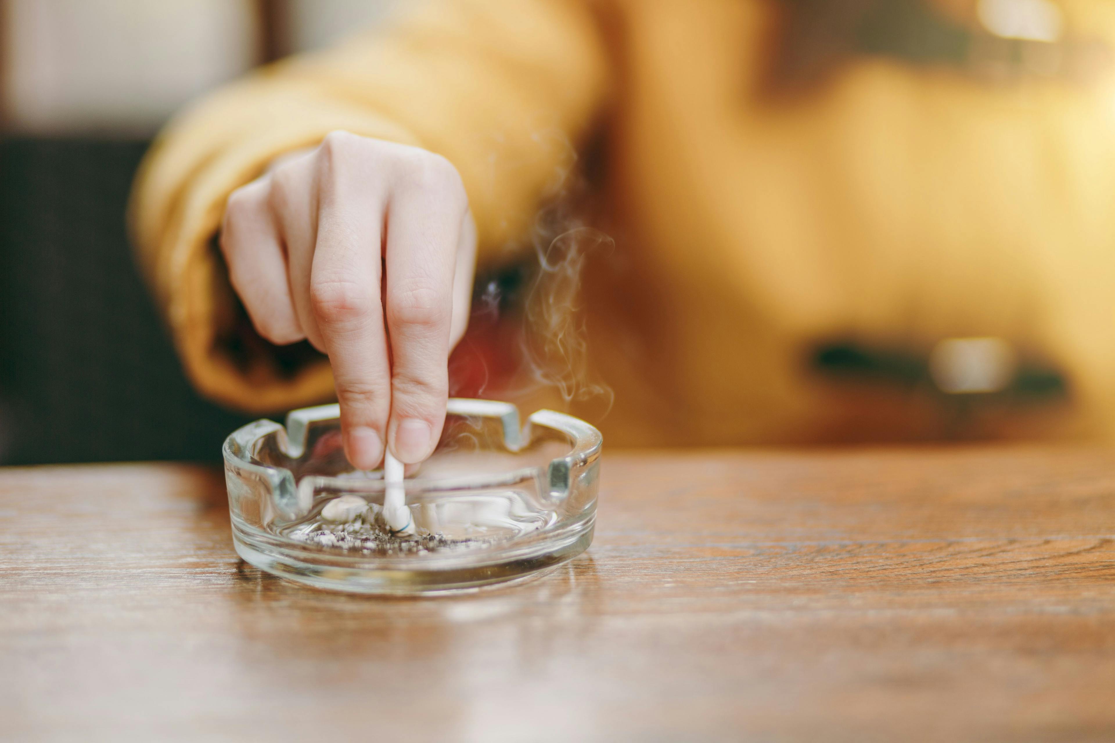Focus on caucasian young woman hand putting out cigarette on glass ashtray on wooden table, cigarette butt, smoking is dying. Quit smoking. Health concept. Close up photo. | Image credit: ViDi Studio - © - stock.adobe.com 