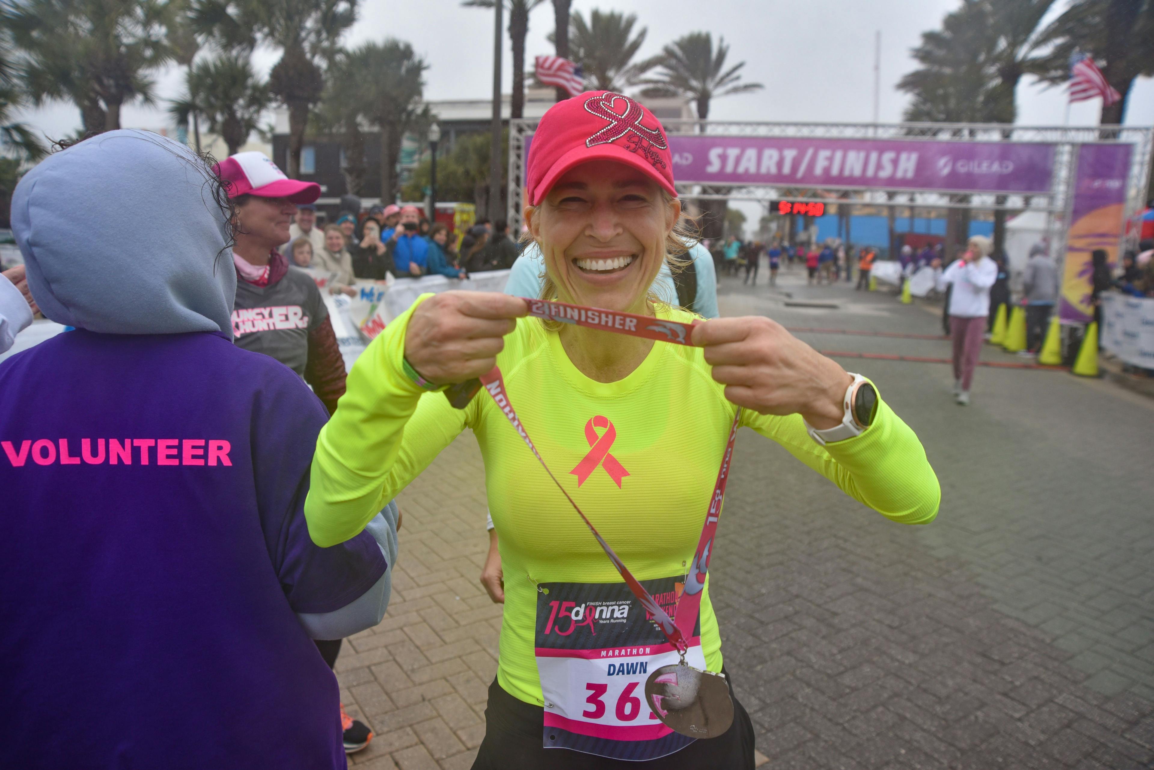Dr. Dawn Mussallem in a bright yellow shirt and pink hat, both with breast cancer ribbons on it, showing off her medal at the finish line of The DONNA Foundaton marathon | Photo credit: The DONNA Foundation