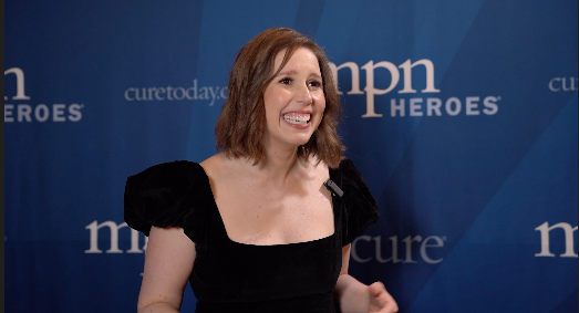 Vanessa Bayer smiling and wearing a black dress standing against a CURE MPN Heroes backdrop