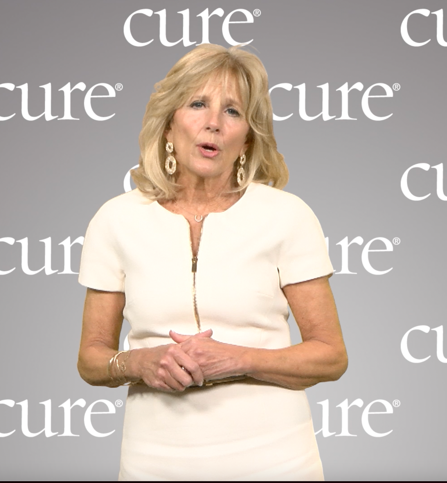 First Lady Jill Biden, in a white dress, speaking in front of a gray background that says "CURE"
