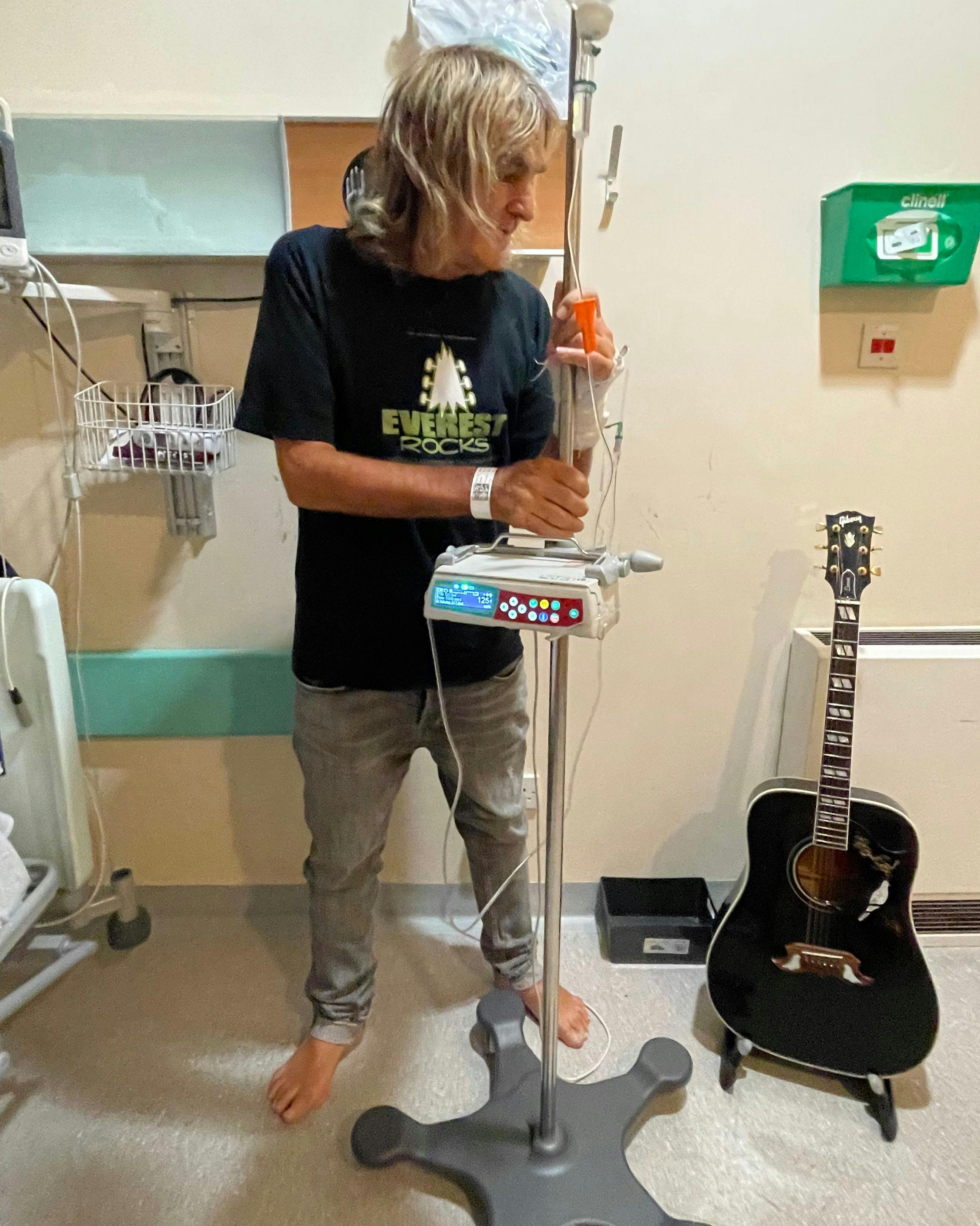 Mike Peters in the hospital holding a black guitar and chemo pole