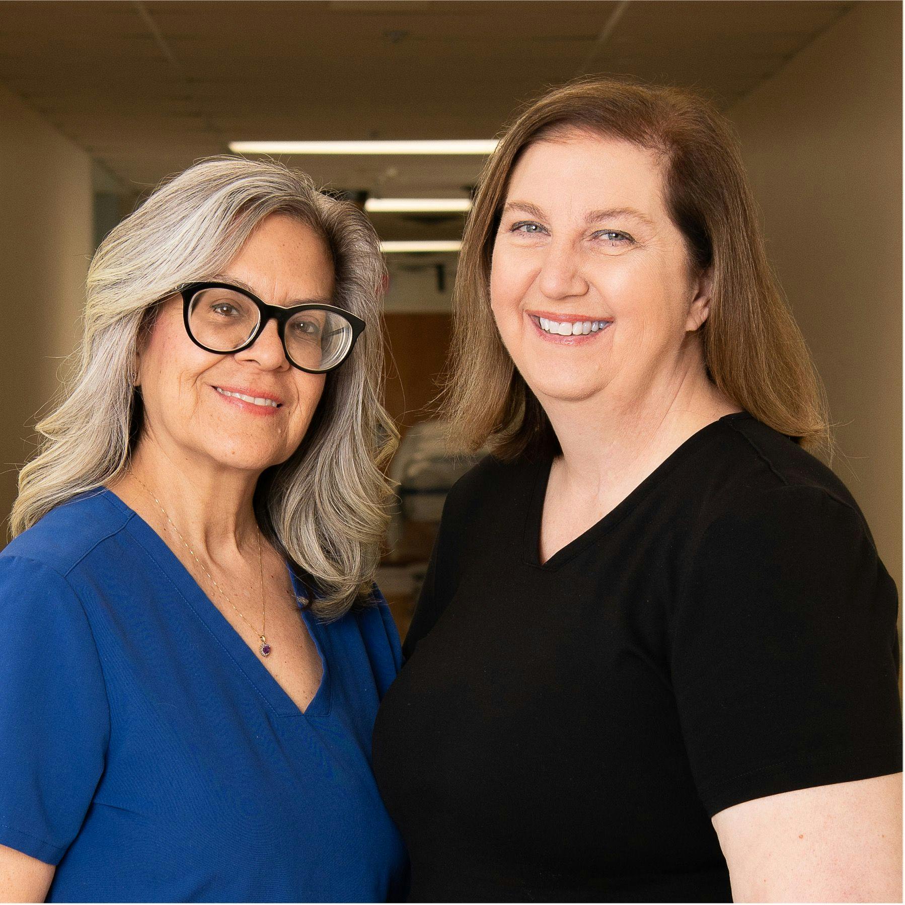 From left: Cynthia Dasaad, M.S.N., RN, OCN, a blonde nurse with blue scrubs and thick black glasses and Deborah Lorick, M.S.N., M.H.A., RN, OCN, a nurse with black scrubs. Both are smiling at the camera.   Photo by Nadia Kelm 