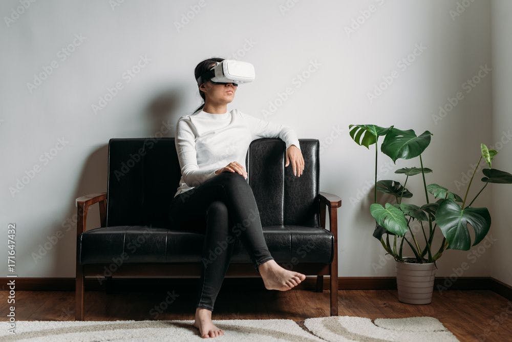 Woman wearing virtual reality headset | Image credit: © - Maa Hoo/Stocksy © - stock.adobe.com. Image of a person sitting on a couch, wearing a virtual reality headset.
