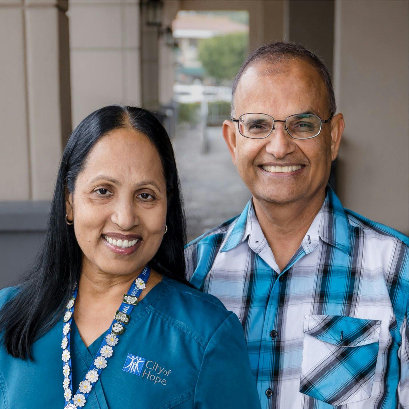 From left: Sobha Akkar, B.S.N., RN, OCN, an oncology nurse in teal scrubs, wearing a lanyard filled with daisy pins and Narayan Ray Akkar, a man in a blue and white plaid shirt, wearing glasses. both are smiling at the camera |   Photos by Alyssa Stefek